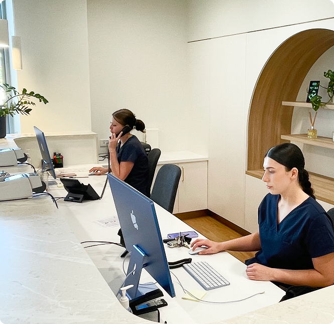 Two dental receptionists in navy scrubs working at a white marble front desk with a modern arched wood shelf.